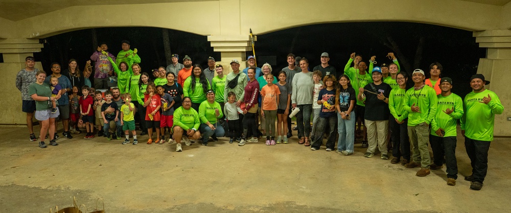 A large group of approximately 50+ people, including adults and children, are posed together for a group photo in what appears to be an outdoor covered pavilion or event space at nighttime. Many participants are wearing matching neon green shirts, suggesting an organized group or volunteer team. The gathering appears to be a community event or volunteer activity, with participants holding small items and displaying celebratory gestures.