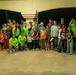 A large group of approximately 50+ people, including adults and children, are posed together for a group photo in what appears to be an outdoor covered pavilion or event space at nighttime. Many participants are wearing matching neon green shirts, suggesting an organized group or volunteer team. The gathering appears to be a community event or volunteer activity, with participants holding small items and displaying celebratory gestures.