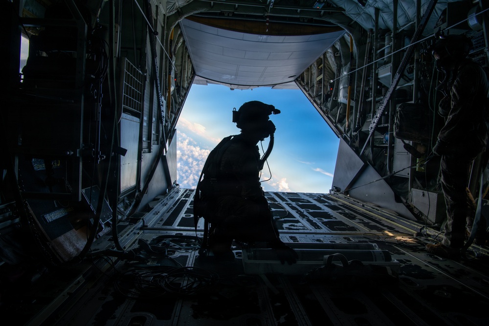 A silhouetted military personnel stands at the open rear cargo ramp of a military transport aircraft in flight, looking out toward a blue sky with clouds below. The figure is equipped with a helmet, communications headset, and tactical gear. The interior of the aircraft cargo bay is visible, including roller tracks on the ramp floor and structural rigging.