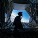 A silhouetted military personnel stands at the open rear cargo ramp of a military transport aircraft in flight, looking out toward a blue sky with clouds below. The figure is equipped with a helmet, communications headset, and tactical gear. The interior of the aircraft cargo bay is visible, including roller tracks on the ramp floor and structural rigging.