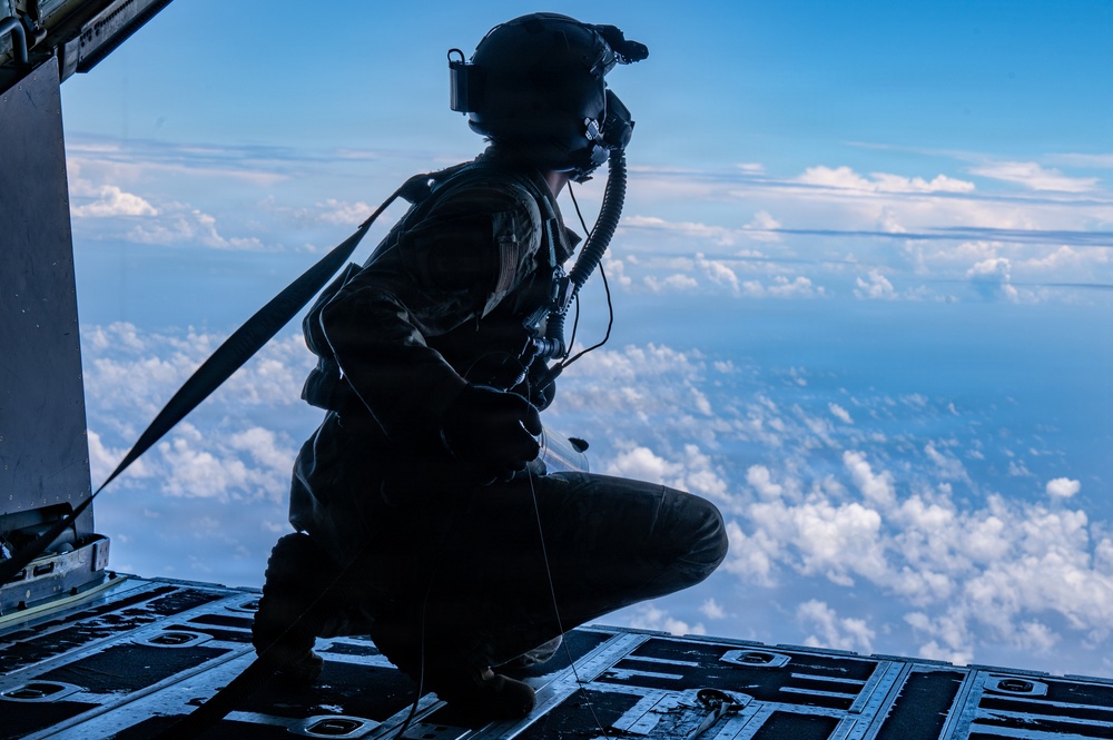 A military aircrew member is crouching on the open rear ramp of a large military transport aircraft at high altitude. The figure is silhouetted against a bright blue sky filled with scattered clouds far below. The person is wearing full flight gear including a helmet with attached equipment, oxygen mask, and harness.