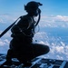 A military aircrew member is crouching on the open rear ramp of a large military transport aircraft at high altitude. The figure is silhouetted against a bright blue sky filled with scattered clouds far below. The person is wearing full flight gear including a helmet with attached equipment, oxygen mask, and harness.