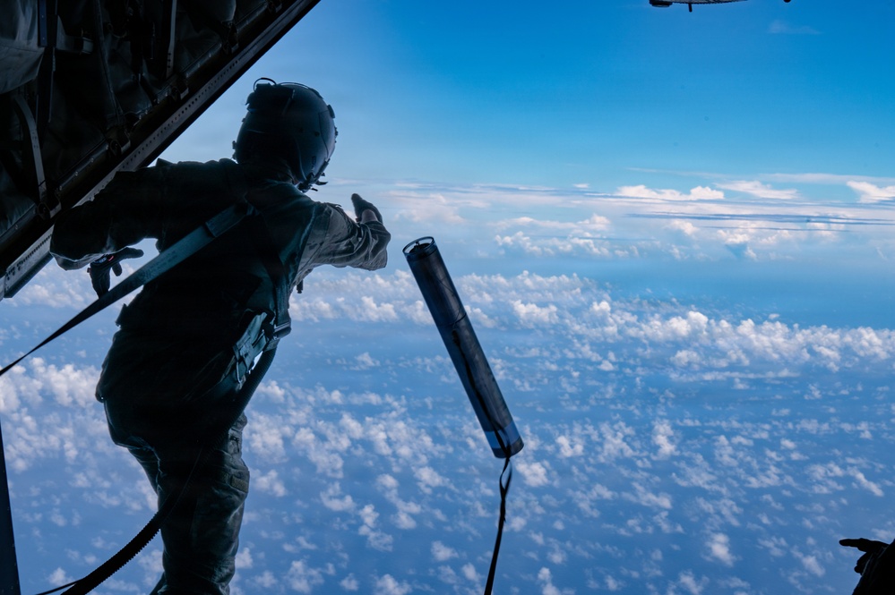 A military aircrew member stands at the open rear ramp of a large aircraft at high altitude, deploying a cylindrical instrument or dropsonde into the atmosphere. The person is in full flight gear including a helmet and is tethered to the aircraft for safety. The scene is set against a backdrop of blue sky and a dense layer of cumulus clouds below.