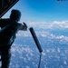 A military aircrew member stands at the open rear ramp of a large aircraft at high altitude, deploying a cylindrical instrument or dropsonde into the atmosphere. The person is in full flight gear including a helmet and is tethered to the aircraft for safety. The scene is set against a backdrop of blue sky and a dense layer of cumulus clouds below.