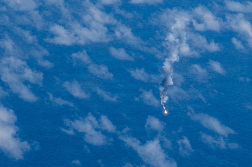 An aerial or orbital photograph showing a rocket or missile in flight, captured from a high altitude perspective over an ocean or large body of water. A bright flame or exhaust plume is visible at the base of the object, with a white smoke trail extending upward and dispersing into a larger cloud formation. The image appears to have been taken from an aircraft or spacecraft looking downward.