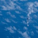 An aerial or orbital photograph showing a rocket or missile in flight, captured from a high altitude perspective over an ocean or large body of water. A bright flame or exhaust plume is visible at the base of the object, with a white smoke trail extending upward and dispersing into a larger cloud formation. The image appears to have been taken from an aircraft or spacecraft looking downward.