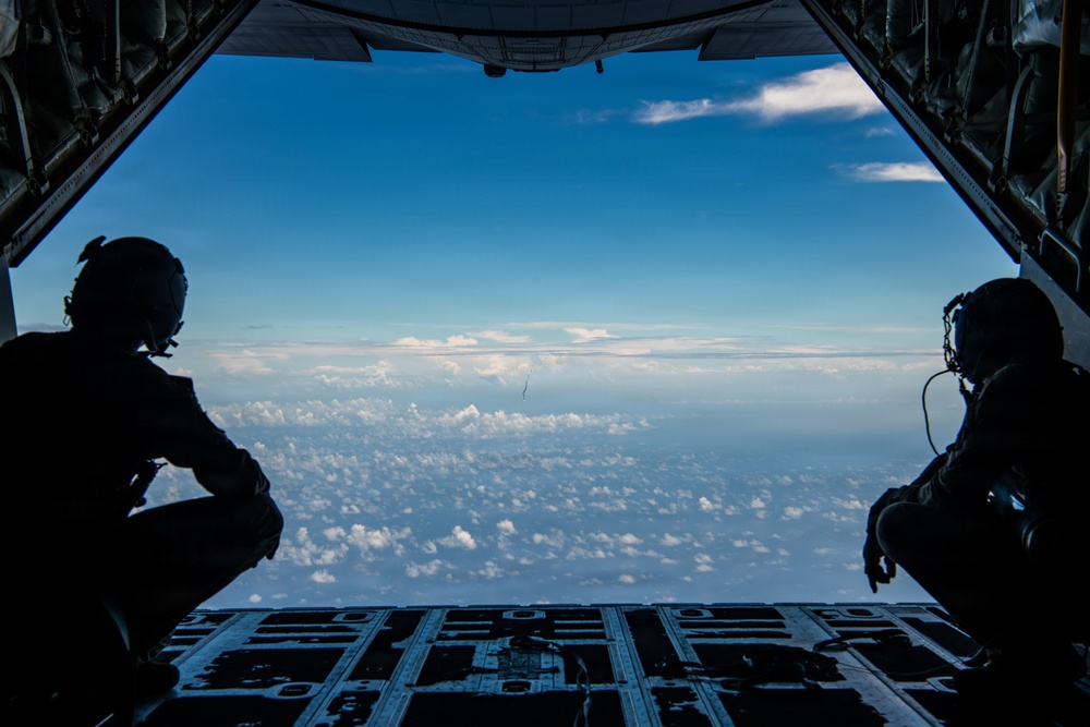 The image is taken from inside a military cargo aircraft with its rear ramp open, showing two silhouetted crew members seated on either side of the open ramp. The view looks out over a vast sky with scattered cumulus clouds at altitude. The scene suggests an airborne operation or observation mission in progress.