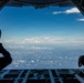 The image is taken from inside a military cargo aircraft with its rear ramp open, showing two silhouetted crew members seated on either side of the open ramp. The view looks out over a vast sky with scattered cumulus clouds at altitude. The scene suggests an airborne operation or observation mission in progress.