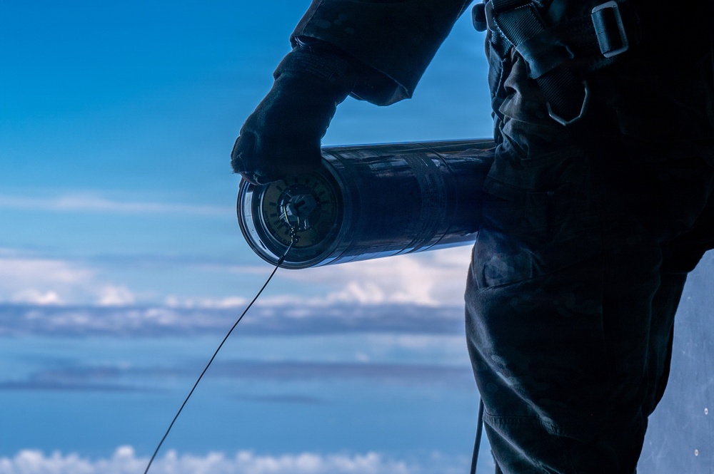 A person dressed in dark tactical/military clothing and gloves is holding a cylindrical device with a wire or line extending from it, photographed against a high-altitude sky with clouds visible below. The perspective and cloud layer suggest the individual is at significant elevation, possibly aboard an aircraft or elevated platform. The image is dramatically lit with the subject appearing largely in silhouette against a blue sky.