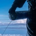 A person dressed in dark tactical/military clothing and gloves is holding a cylindrical device with a wire or line extending from it, photographed against a high-altitude sky with clouds visible below. The perspective and cloud layer suggest the individual is at significant elevation, possibly aboard an aircraft or elevated platform. The image is dramatically lit with the subject appearing largely in silhouette against a blue sky.