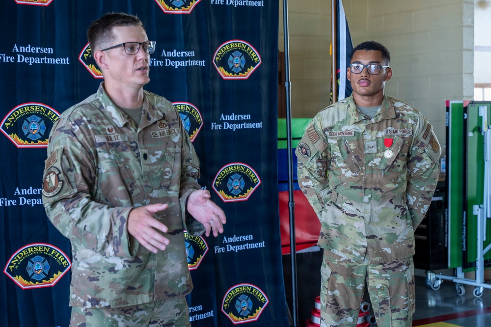 Two U.S. Air Force personnel are standing in front of an Andersen Fire Department backdrop, likely during a formal recognition ceremony. The airman on the right is wearing a red medal on his uniform, suggesting he is being honored or has recently received an award. The senior airman on the left appears to be speaking or presenting.