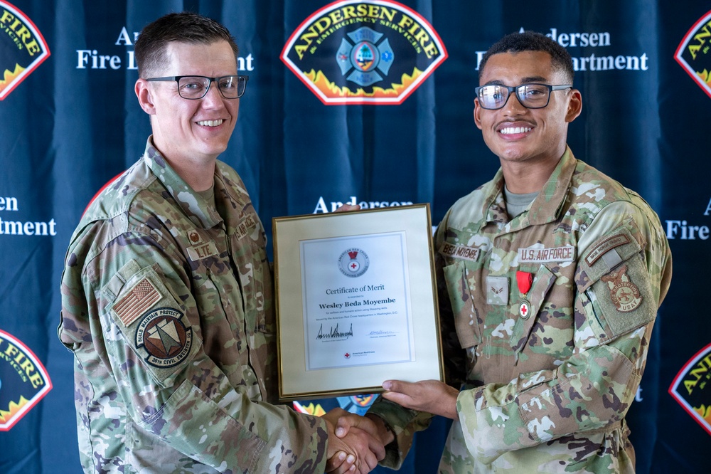 Two U.S. Air Force personnel in MultiCam/OCP uniforms are shaking hands while jointly holding a framed American Red Cross Certificate of Merit awarded to Wesley Beda Moyembe. The ceremony is taking place in front of an Andersen Fire Department backdrop, indicating this is at Andersen Air Force Base. The recipient on the right is wearing a red medal on his uniform.