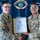 Two U.S. Air Force personnel in MultiCam/OCP uniforms are shaking hands while jointly holding a framed American Red Cross Certificate of Merit awarded to Wesley Beda Moyembe. The ceremony is taking place in front of an Andersen Fire Department backdrop, indicating this is at Andersen Air Force Base. The recipient on the right is wearing a red medal on his uniform.