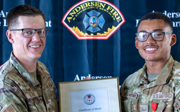 Two U.S. Air Force personnel in MultiCam/OCP uniforms are shaking hands while jointly holding a framed American Red Cross Certificate of Merit awarded to Wesley Beda Moyembe. The ceremony is taking place in front of an Andersen Fire Department backdrop, indicating this is at Andersen Air Force Base. The recipient on the right is wearing a red medal on his uniform.