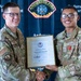 Two U.S. Air Force personnel in MultiCam/OCP uniforms are shaking hands while jointly holding a framed American Red Cross Certificate of Merit awarded to Wesley Beda Moyembe. The ceremony is taking place in front of an Andersen Fire Department backdrop, indicating this is at Andersen Air Force Base. The recipient on the right is wearing a red medal on his uniform.