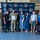 A group of approximately ten people pose for a formal photo in front of an Andersen Fire Department backdrop, likely at a recognition or award ceremony. A military service member in camouflage uniform is holding a framed certificate or award, suggesting this is an official commendation event. The group includes military personnel, civilians, and a service dog.