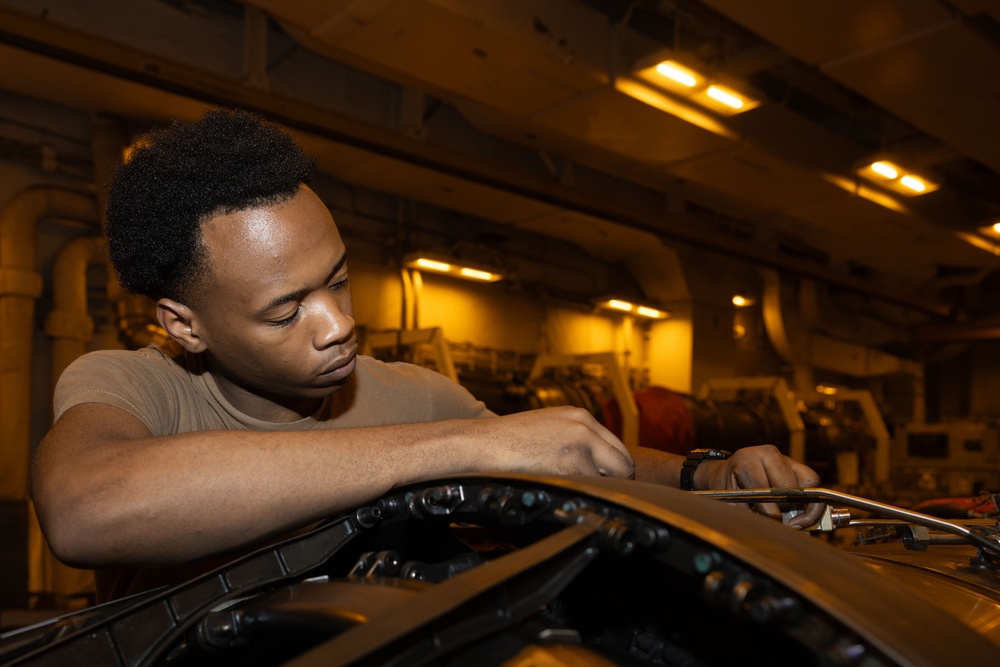 A young man in a tan/olive military-style t-shirt is using a ratchet wrench to perform maintenance work on what appears to be a military vehicle or aircraft component. The setting appears to be an enclosed hangar or ship hangar bay, illuminated by warm amber overhead lighting. The worker is focused and leaning over the equipment with concentrated attention.