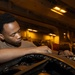 A young man in a tan/olive military-style t-shirt is using a ratchet wrench to perform maintenance work on what appears to be a military vehicle or aircraft component. The setting appears to be an enclosed hangar or ship hangar bay, illuminated by warm amber overhead lighting. The worker is focused and leaning over the equipment with concentrated attention.