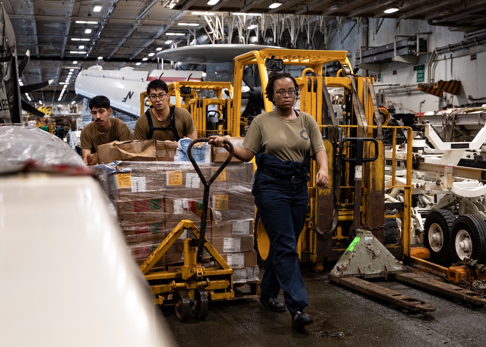 Three military personnel in olive/khaki uniforms are moving a large pallet of supplies using a manual pallet jack inside what appears to be the hangar bay of a naval aircraft carrier. The space contains multiple aircraft and yellow ground support equipment. The workers appear to be conducting a replenishment or logistics operation.