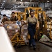 Three military personnel in olive/khaki uniforms are moving a large pallet of supplies using a manual pallet jack inside what appears to be the hangar bay of a naval aircraft carrier. The space contains multiple aircraft and yellow ground support equipment. The workers appear to be conducting a replenishment or logistics operation.