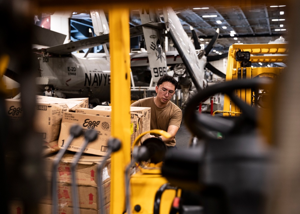 A U.S. Navy sailor in a tan uniform is handling cardboard boxes labeled 'Eggo' on a pallet inside what appears to be an aircraft carrier hangar bay. Yellow forklifts and material handling equipment are visible in the foreground, while a Navy E-2D Hawkeye aircraft with tail number 986 and markings '601' is visible in the background. The scene depicts routine logistics and supply operations aboard a naval vessel.