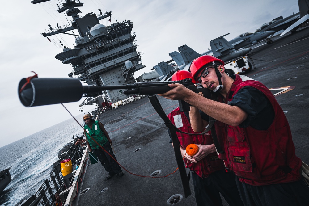 Personnel in red vests and red hard hats are operating what appears to be a line-throwing gun on the flight deck of a large aircraft carrier. The foreground sailor is aiming the device while a second person holds an orange line spool. A third crew member in a green vest observes from the port side edge of the deck.