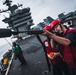 Personnel in red vests and red hard hats are operating what appears to be a line-throwing gun on the flight deck of a large aircraft carrier. The foreground sailor is aiming the device while a second person holds an orange line spool. A third crew member in a green vest observes from the port side edge of the deck.