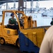 A military naval personnel operates a yellow Hyster forklift (ID: 13-33062) aboard what appears to be a U.S. Navy vessel, likely in a hangar bay or cargo deck area. The forklift is marked 'Shipboard Use Approved' and 'Structurally Tested by Hyster,' with a vehicle weight of 11,360 lbs. Multiple crew members in green safety vests and blue hard hats are visible in the background, with other naval vessels docked nearby.