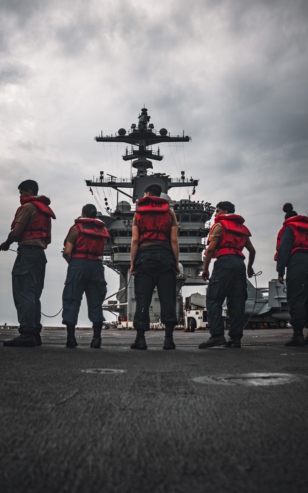Several naval personnel wearing red life vests stand on the flight deck of a large aircraft carrier, facing the ship's island superstructure. The low-angle perspective emphasizes the imposing scale of the carrier's tower and radar/communications equipment against an overcast sky. At least one military aircraft is partially visible on the right side of the deck.