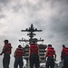 Several naval personnel wearing red life vests stand on the flight deck of a large aircraft carrier, facing the ship's island superstructure. The low-angle perspective emphasizes the imposing scale of the carrier's tower and radar/communications equipment against an overcast sky. At least one military aircraft is partially visible on the right side of the deck.