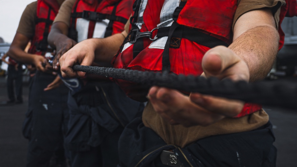 Multiple individuals wearing red life vests and dark pants are gripping a thick black rope, suggesting a coordinated line-handling or mooring operation. The close-up perspective emphasizes their hands and upper torsos, indicating teamwork in a maritime or naval setting. The wet texture on the rope and the overcast background suggest outdoor conditions near water.