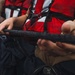 Multiple individuals wearing red life vests and dark pants are gripping a thick black rope, suggesting a coordinated line-handling or mooring operation. The close-up perspective emphasizes their hands and upper torsos, indicating teamwork in a maritime or naval setting. The wet texture on the rope and the overcast background suggest outdoor conditions near water.
