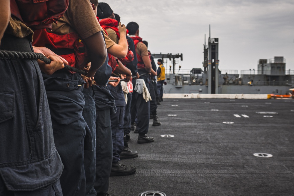 A line of naval personnel standing on the deck of a warship, holding a mooring or tow line during what appears to be an underway replenishment or mooring operation. The crew members are wearing red life vests over their working uniforms. A second vessel is visible in the background, suggesting a ship-to-ship operation.