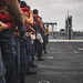 A line of naval personnel standing on the deck of a warship, holding a mooring or tow line during what appears to be an underway replenishment or mooring operation. The crew members are wearing red life vests over their working uniforms. A second vessel is visible in the background, suggesting a ship-to-ship operation.