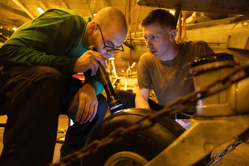 Two individuals are closely inspecting a large mechanical component, likely a wheel or tire assembly, in what appears to be a dimly lit hangar or ship deck bay. One person wearing a green shirt and glasses is using a flashlight to illuminate the area under inspection, while the other in an olive military-style shirt observes closely. The setting and attire suggest a military aviation maintenance context.
