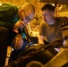 Two individuals are closely inspecting a large mechanical component, likely a wheel or tire assembly, in what appears to be a dimly lit hangar or ship deck bay. One person wearing a green shirt and glasses is using a flashlight to illuminate the area under inspection, while the other in an olive military-style shirt observes closely. The setting and attire suggest a military aviation maintenance context.