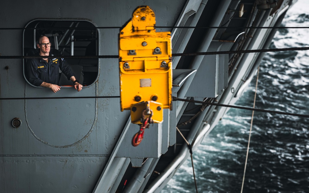 A U.S. Navy sailor leans out of a porthole window on the gray hull of a naval vessel underway at sea. A prominent yellow crane or davit hook assembly is mounted on the ship's exterior in the foreground. The ship appears to be moving through choppy open water, visible in the background.