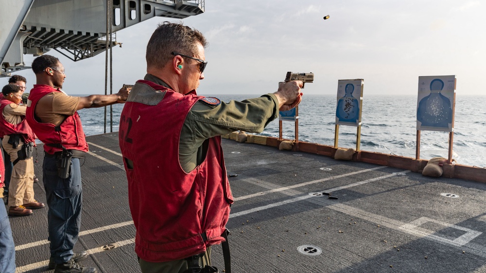 Military or naval personnel are conducting a live-fire pistol qualification exercise on the deck of a naval vessel at sea. A spent brass casing is visibly ejecting from the foreground shooter's pistol, indicating an active firing moment. Multiple silhouette paper targets showing bullet impact patterns are positioned downrange near the ship's railing.