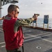 Military or naval personnel are conducting a live-fire pistol qualification exercise on the deck of a naval vessel at sea. A spent brass casing is visibly ejecting from the foreground shooter's pistol, indicating an active firing moment. Multiple silhouette paper targets showing bullet impact patterns are positioned downrange near the ship's railing.