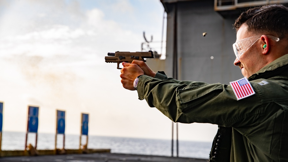 A U.S. military service member is conducting a live-fire pistol qualification exercise on the deck of a naval vessel at sea. The shooter is firing a semi-automatic pistol toward a range of blue target markers, with a spent brass casing visibly ejecting from the firearm. The setting appears to be an outdoor shipboard firing range during daylight hours.