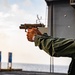 A U.S. military service member is conducting a live-fire pistol qualification exercise on the deck of a naval vessel at sea. The shooter is firing a semi-automatic pistol toward a range of blue target markers, with a spent brass casing visibly ejecting from the firearm. The setting appears to be an outdoor shipboard firing range during daylight hours.