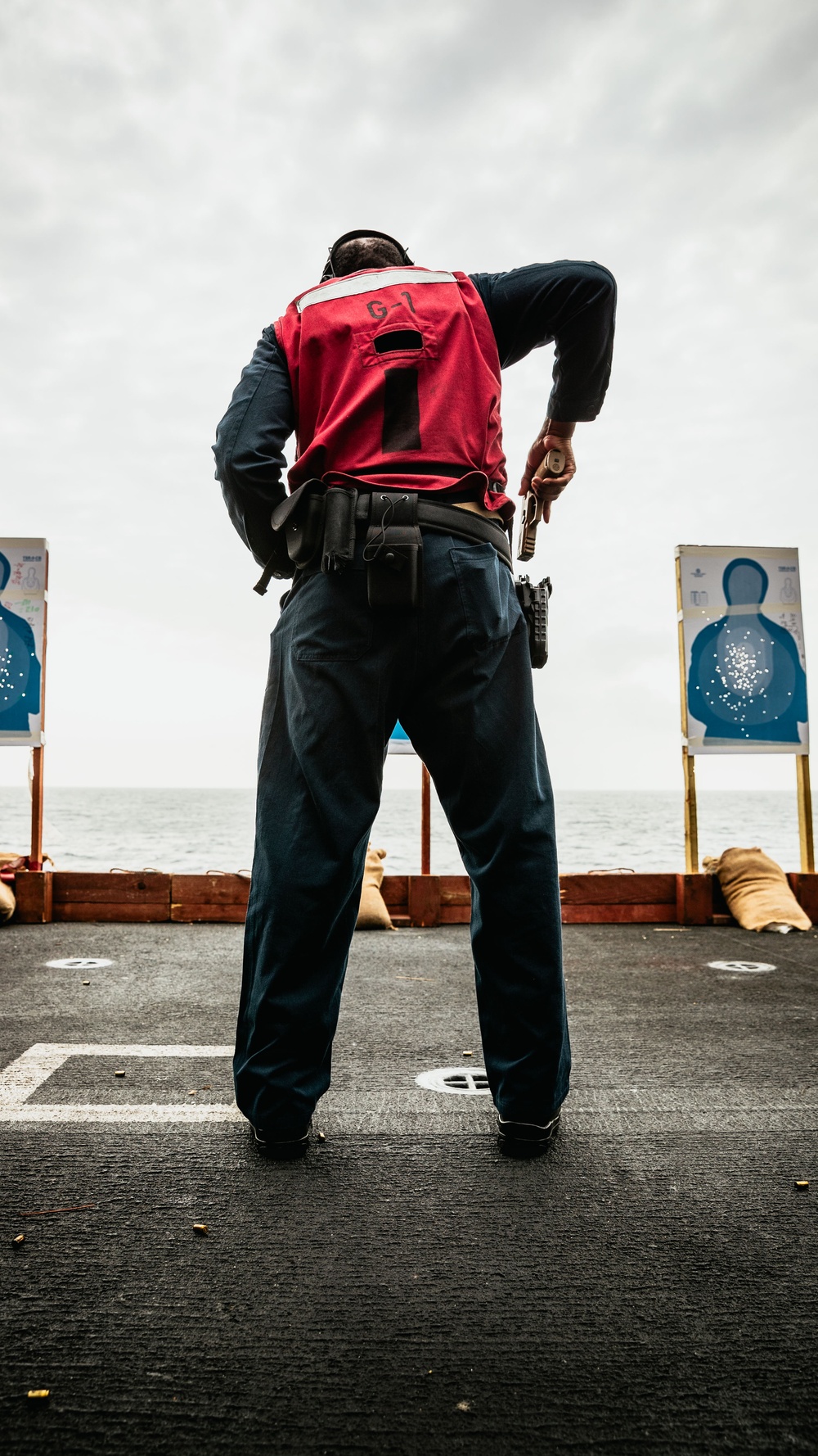 A person wearing a red vest marked 'G-1' stands on what appears to be the deck of a naval vessel, holding a pistol at their side while facing two blue silhouette shooting targets. The setting is an outdoor firearms qualification range set up on a ship's deck, with the ocean visible in the background. Sandbags and wooden barriers are positioned behind the targets as a backstop.