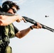 A person is actively firing a pump-action shotgun at what appears to be an outdoor shooting range. A spent shell casing is visibly ejecting from the firearm, captured mid-air to the right of the shooter. The individual is wearing appropriate hearing and eye protection.