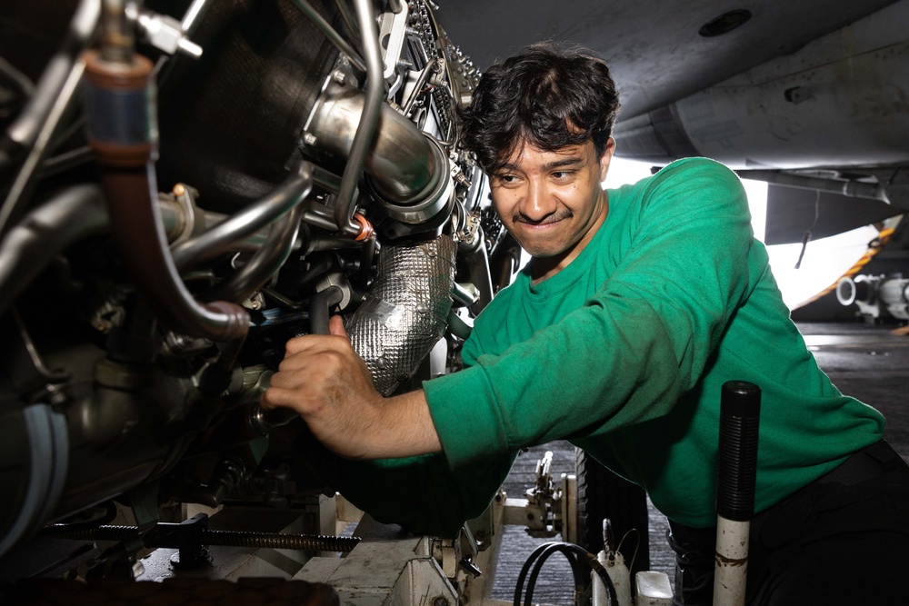 A young male technician wearing a green shirt is performing maintenance work on an aircraft engine in what appears to be a hangar bay or flight deck environment. He is using a hand tool to work on engine components and appears focused and slightly smiling. The setting suggests a military or naval aviation maintenance context.