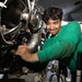 A young male technician wearing a green shirt is performing maintenance work on an aircraft engine in what appears to be a hangar bay or flight deck environment. He is using a hand tool to work on engine components and appears focused and slightly smiling. The setting suggests a military or naval aviation maintenance context.