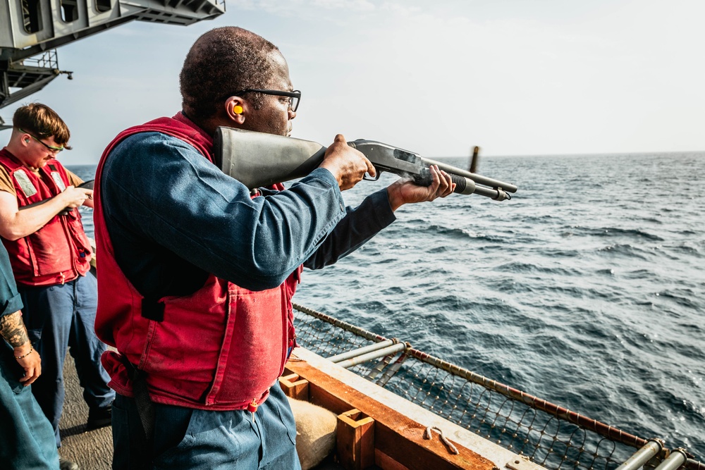 A person wearing a red life vest and orange ear protection is aiming a pump-action shotgun over the railing of a naval vessel toward open ocean. At least two other individuals in similar red life vests are visible in the background on the ship's deck. The setting appears to be a military or coast guard vessel conducting a live-fire exercise at sea.