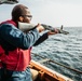 A person wearing a red life vest and orange ear protection is aiming a pump-action shotgun over the railing of a naval vessel toward open ocean. At least two other individuals in similar red life vests are visible in the background on the ship's deck. The setting appears to be a military or coast guard vessel conducting a live-fire exercise at sea.