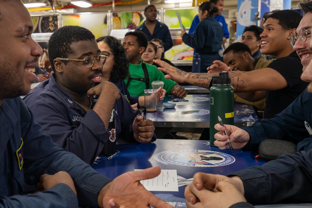 A group of military personnel, likely U.S. Navy sailors, are gathered in what appears to be a ship's mess hall or dining facility. They are engaged in animated conversation and appear to be in a relaxed, social setting. The table features a decorative surface with what appears to be a military or naval insignia/seal.