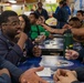 A group of military personnel, likely U.S. Navy sailors, are gathered in what appears to be a ship's mess hall or dining facility. They are engaged in animated conversation and appear to be in a relaxed, social setting. The table features a decorative surface with what appears to be a military or naval insignia/seal.