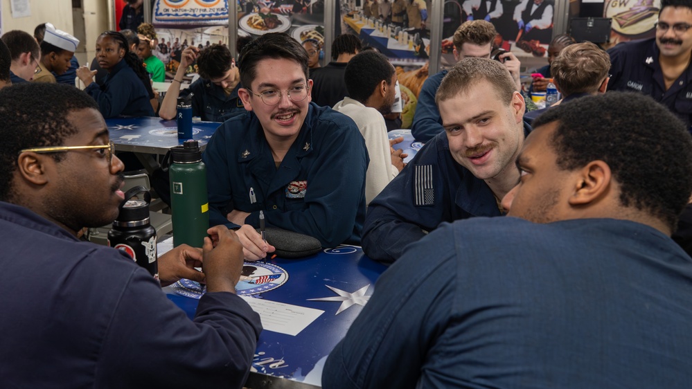 A group of military personnel in navy blue uniforms are gathered around a table in what appears to be a mess hall or dining facility aboard a naval vessel. Several sailors are engaged in conversation, smiling and interacting in a relaxed social setting. The background shows additional personnel seated at other tables in a busy dining environment.